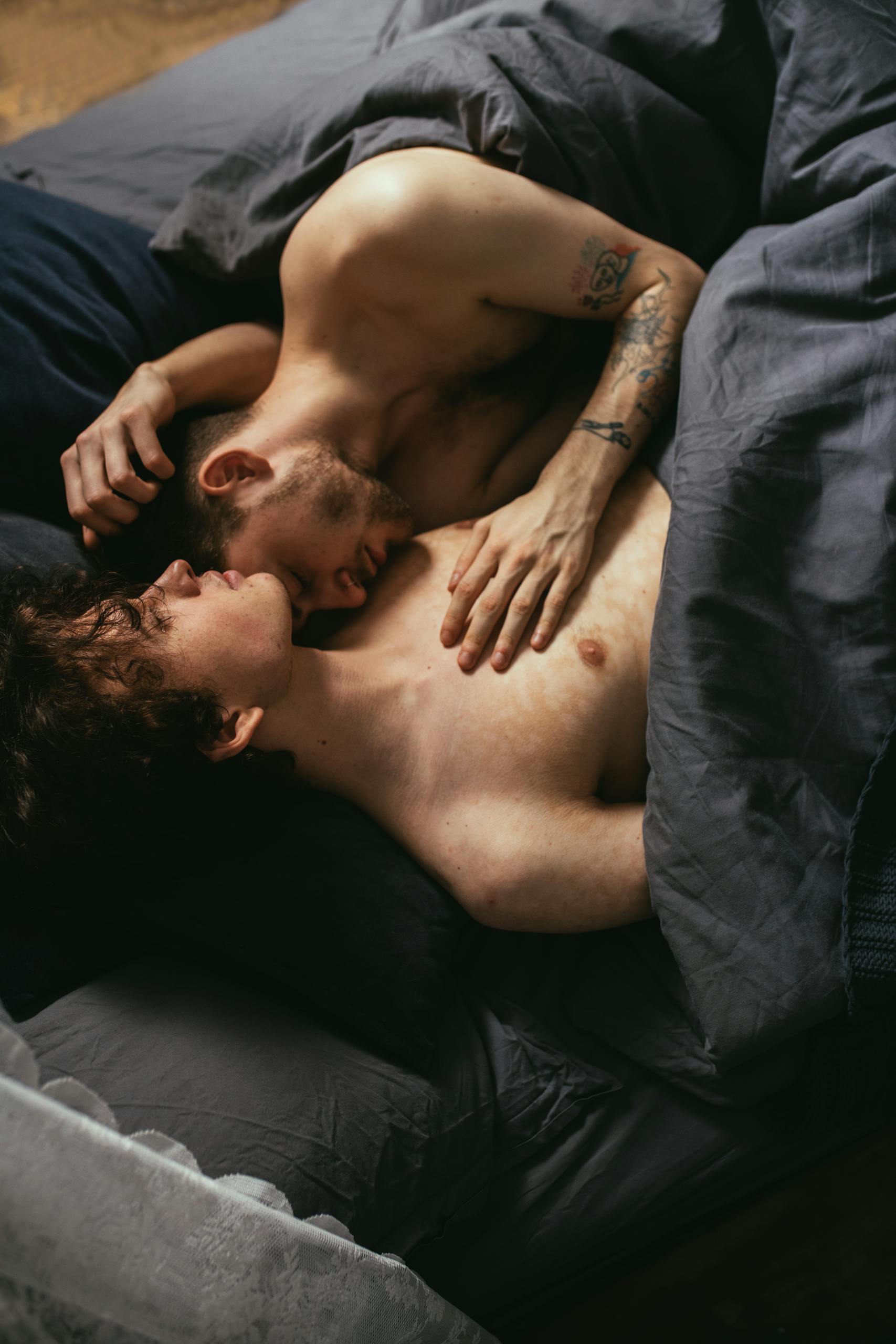 Tender moment of a shirtless gay couple lying in bed, embracing each other lovingly.