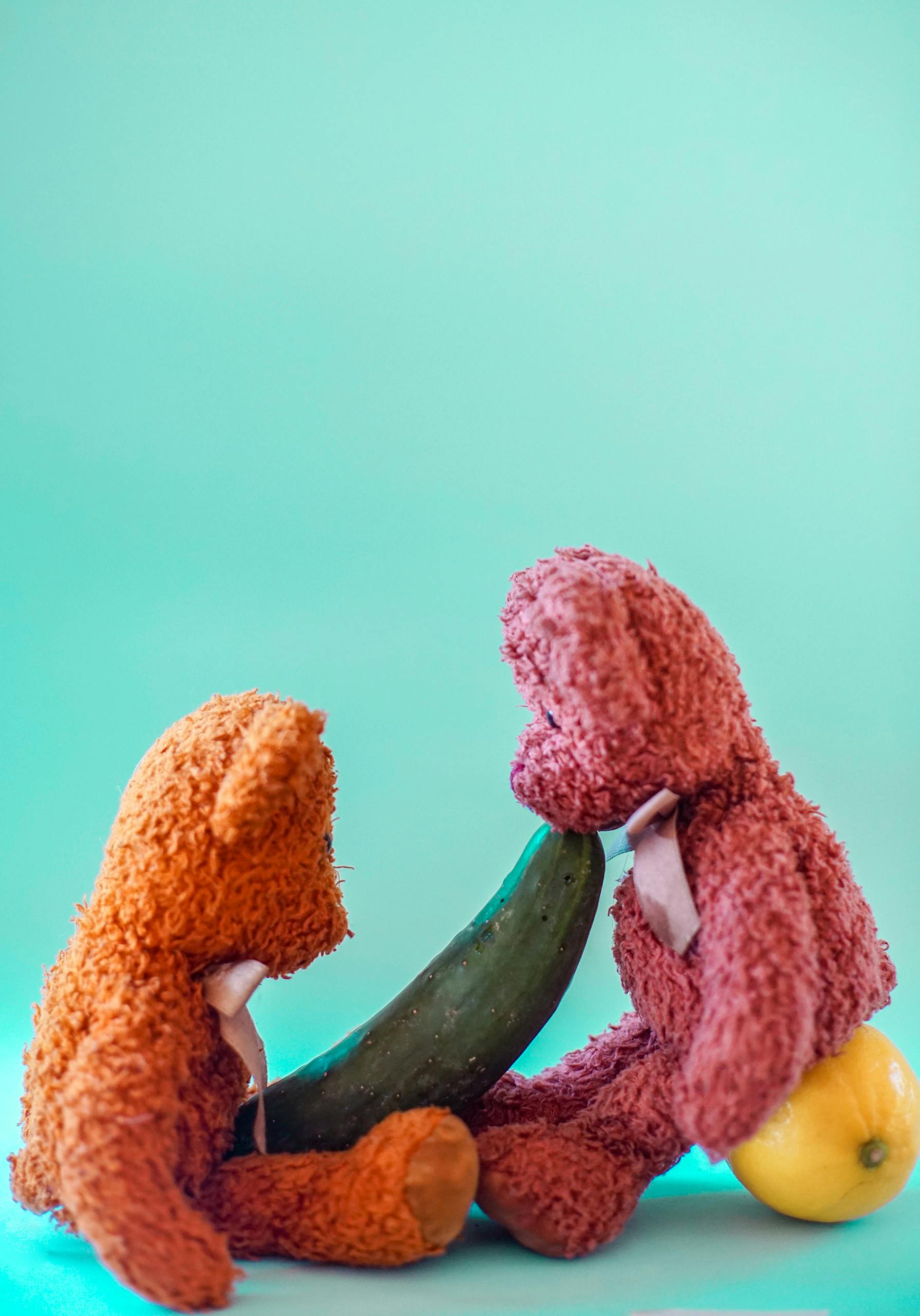 Two colorful teddy bears interacting creatively with fruits and cucumber.